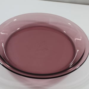 May include: A round, transparent, purple glass pie dish. The dish has a slightly raised rim and a flat bottom. The color is a deep, rich purple, and the glass appears to be smooth and clear. The dish is empty and sits on a white surface.