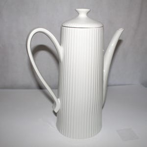May include: A tall, white ceramic coffee pot with a lid and a curved handle. The pot has vertical ribbed detailing and a long, slender spout. The design is minimalist and elegant, suitable for serving coffee or tea.