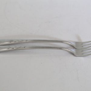 May include: Two stainless steel forks with a simple, elegant design. The forks have a classic shape with three tines and a long, slender handle. The handles feature a subtle decorative pattern. The forks are set against a plain white background.