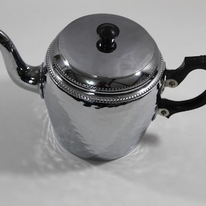 May include: A shiny, silver-colored teapot with a black handle and lid knob. The teapot has a hammered texture and a decorative beaded trim around the lid. The spout is curved, and the handle is a curved, open design.
