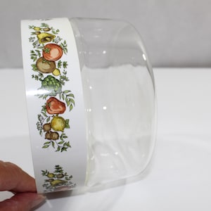 May include: A clear glass bowl with a white band featuring a colorful illustration of various vegetables, including tomatoes, artichokes, and lemons. The bowl is partially visible, showcasing its curved shape and transparent design.