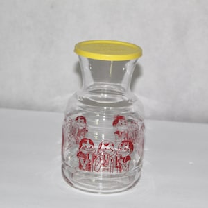 May include: Clear glass pitcher with a yellow lid. The pitcher has a flared neck and a rounded base. Red cartoon figures are printed around the base of the pitcher. The pitcher is likely used for serving beverages.