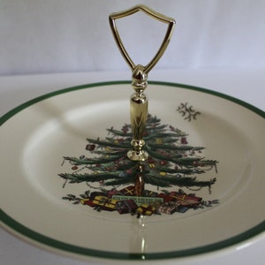 May include: A two-tiered serving plate with a gold handle. The plate features a Christmas tree design with ornaments and wrapped gifts. The rim has a green border, perfect for holiday gatherings.