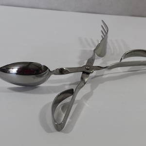 May include: Stainless steel serving tongs with a spoon on one end and a fork on the other. The handles have a looped design for easy gripping. The tongs are designed for serving food.