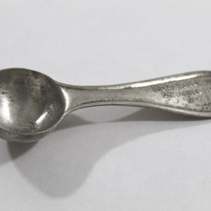 May include: A small, vintage silver-colored spoon with a rounded bowl and a short handle. The handle has embossed text. The spoon appears to be made of metal and shows signs of wear, suggesting it is an antique or a well-used item.