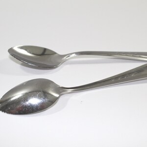 May include: Two stainless steel grapefruit spoons are displayed against a white background. The spoons have serrated edges and decorative detailing on the handles. The spoons are designed for eating grapefruit.