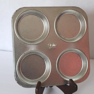 May include: A square, silver-colored muffin tin with four circular molds. The tin has a slightly worn appearance, suggesting it's been used. The brand name "Skyline" is visible. The tin is displayed on a wooden stand.