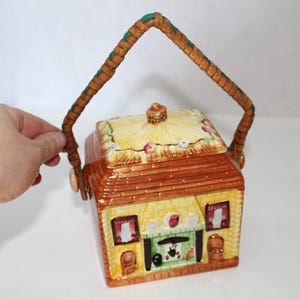 May include: A ceramic house-shaped cookie jar with a woven handle. The jar features a yellow brick facade, a brown roof, and a decorative fireplace. The lid is adorned with floral accents. The handle is brown with green accents.