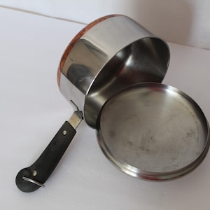 May include: A stainless steel saucepan with a black handle and a lid. The pan has a wooden rim and is angled towards the viewer, with the lid slightly ajar. The kitchenware is on a white surface.