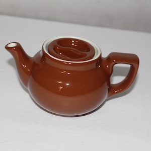 May include: A small, brown ceramic teapot with a white rim and a matching lid. The teapot has a curved spout and a handle. The design is simple and classic, suitable for serving tea. The teapot is approximately 5 inches long.