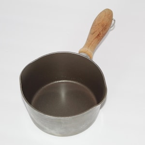 May include: A small, vintage metal saucepan with a wooden handle. The pan has a dark interior and a light-colored exterior. The wooden handle is light brown and attached with a metal ring.