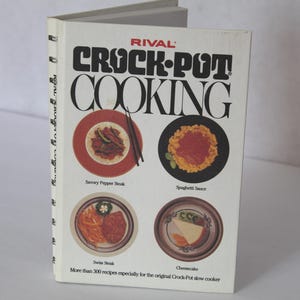 May include: A cookbook titled "Crock-Pot Cooking" by Rival. The cover features the title in large black letters with images of prepared dishes, including savory pepper steak, spaghetti sauce, Swiss steak, and cheesecake. The book is open.