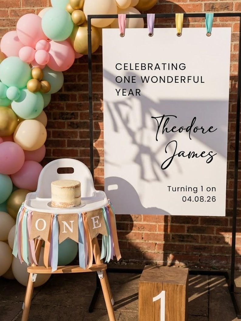May include: A white high chair with a burlap banner reading ONE, decorated with pastel ribbons. Beside it, a white sign on a black frame reads CELEBRATING ONE WONDERFUL YEAR, Theodore James, Turning 1 on 04.08.26, next to a balloon arch and a wooden block with 1.