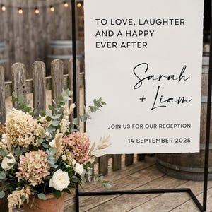 May include: A rectangular white wedding sign with deckled edges, suspended by sage green ribbons from a black metal frame. The text reads: TO LOVE, LAUGHTER AND A HAPPY EVER AFTER, Sarah + Liam, JOIN US FOR OUR RECEPTION, 14 SEPTEMBER 2025.