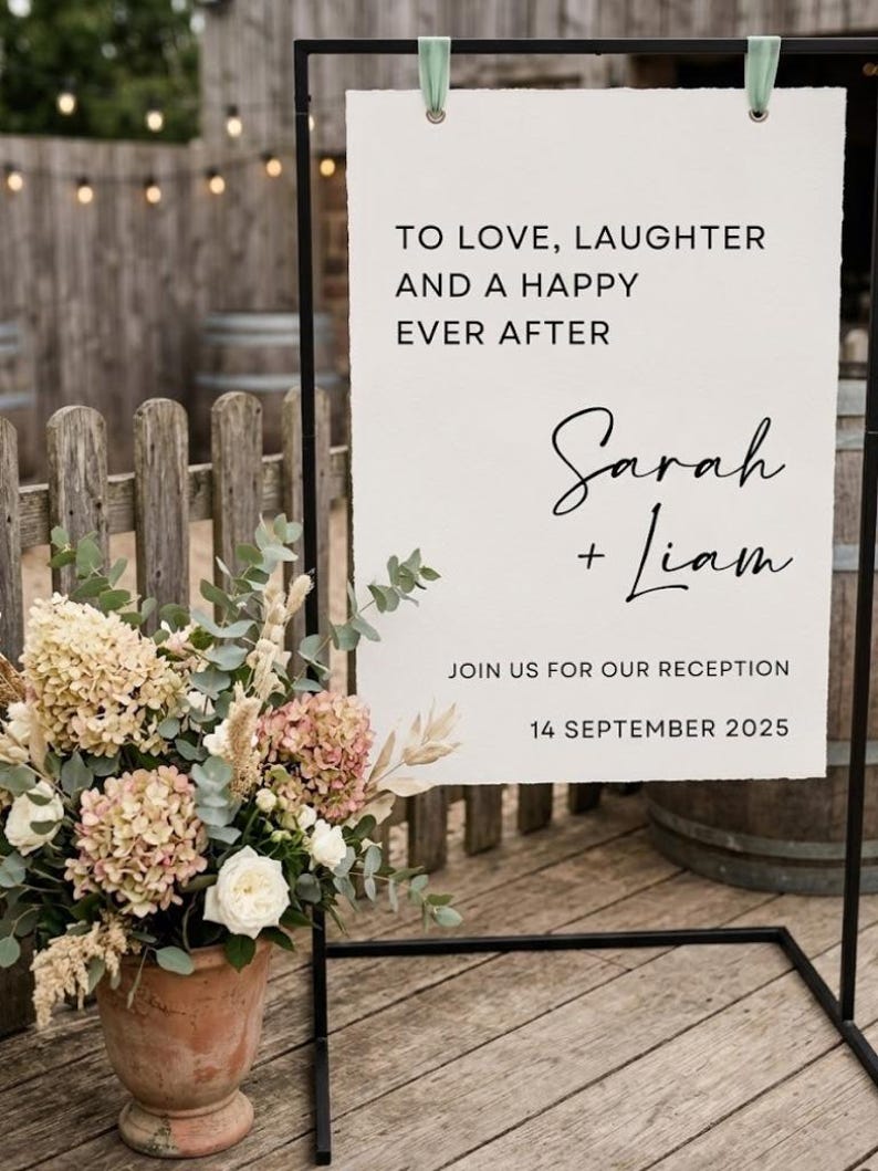May include: A rectangular white wedding sign with deckled edges, suspended by sage green ribbons from a black metal frame. The text reads: TO LOVE, LAUGHTER AND A HAPPY EVER AFTER, Sarah + Liam, JOIN US FOR OUR RECEPTION, 14 SEPTEMBER 2025.