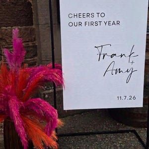May include: A white rectangular sign suspended from a black metal frame, featuring the black text "CHEERS TO OUR FIRST YEAR", "Frank + Amy", and "11.7.26". Beside the sign is a tall, dark brown vase filled with vibrant, bright pink and orange pampas grass.