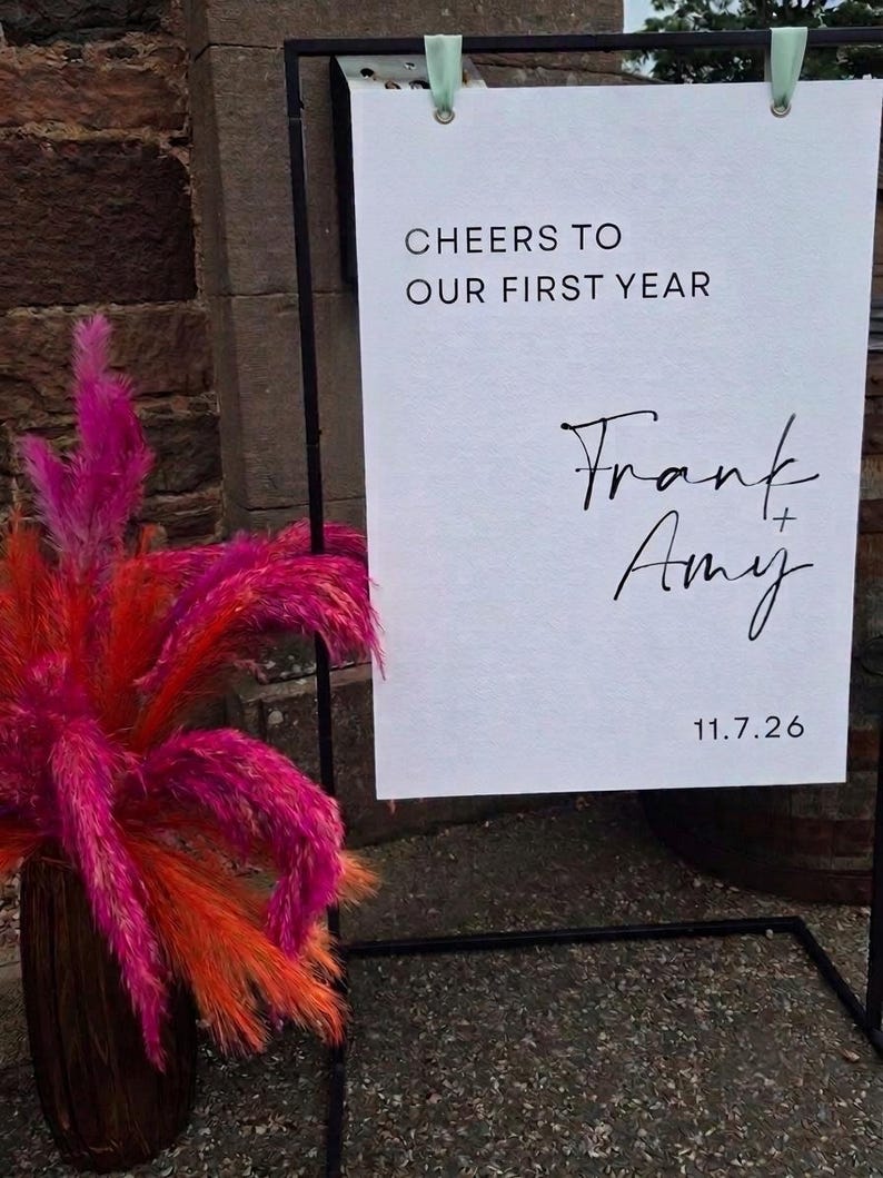 May include: A white rectangular sign suspended from a black metal frame, featuring the black text "CHEERS TO OUR FIRST YEAR", "Frank + Amy", and "11.7.26". Beside the sign is a tall, dark brown vase filled with vibrant, bright pink and orange pampas grass.