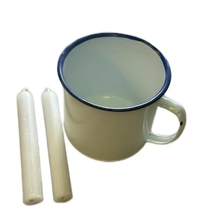 May include: A white enamel mug with a blue rim and handle, accompanied by two white taper candles. The mug has a classic design, suitable for camping or rustic decor. The candles are slender and cylindrical.