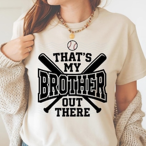 May include: A beige t-shirt with the text "THAT'S MY BROTHER OUT THERE" in a black, bold font, with crossed baseball bats and a baseball graphic. The shirt is a casual style.