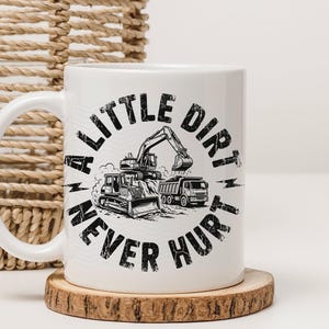 May include: White ceramic mug with a black and white graphic of construction vehicles, including an excavator, bulldozer, and dump truck. The text "A LITTLE DIRT NEVER HURT" is printed around the vehicles. The mug sits on a wooden coaster.