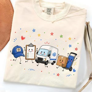 May include: A cream-coloured t-shirt with a Comfort Colors label featuring a graphic of a blue baseball cap, a clipboard, a white delivery van, and a brown cardboard box on a hand truck, all with faces, surrounded by colourful stars and dots.