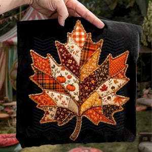 May include: A black fabric square featuring a large, central maple leaf applique composed of various patchwork fabric segments. The leaf displays autumn-themed patterns including orange plaid, floral prints, polka dots, and small pumpkin motifs.