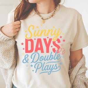 May include: Cream-colored t-shirt with the text "Sunny Days & Double Plays" in yellow, red, and blue. The design includes stars and a baseball graphic. A casual, everyday tee.