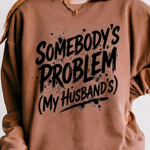 May include: A brown sweatshirt with the text "SOMEBODY'S PROBLEM (MY HUSBAND'S)" in black, with a distressed font and splatter effect. The design includes a gray zigzag pattern.