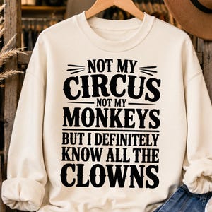 May include: A cream-colored crewneck sweatshirt featuring black text that reads: NOT MY CIRCUS NOT MY MONKEYS BUT I DEFINITELY KNOW ALL THE CLOWNS. The garment has long sleeves with ribbed cuffs and a ribbed neckline, presented on a wooden hanger.