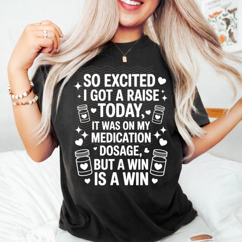 K&ouml;nnte beinhalten: Schwarzes T-Shirt mit wei&szlig;em Text: "SO EXCITED I GOT A RAISE TODAY, IT WAS ON MY MEDICATION DOSAGE, BUT A WIN IS A WIN." Das Design umfasst Pillenflaschen-Grafiken sowie Stern- und Herz-Akzente.