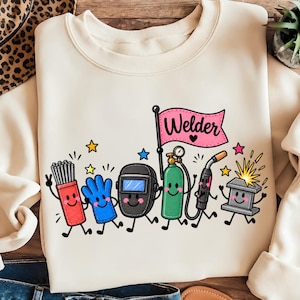 May include: A cream-coloured crewneck sweatshirt featuring a colourful graphic of cartoon welding equipment including a red rod holder, blue glove, black welding helmet, green gas tank, and anvil, with a pink flag labelled Welder with a heart.