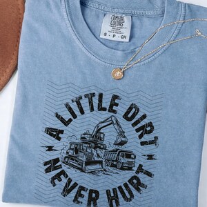 May include: A light blue t-shirt with the text "A Little Dirt Never Hurt" and illustrations of construction vehicles. A gold necklace with a circular pendant is on top of the shirt.
