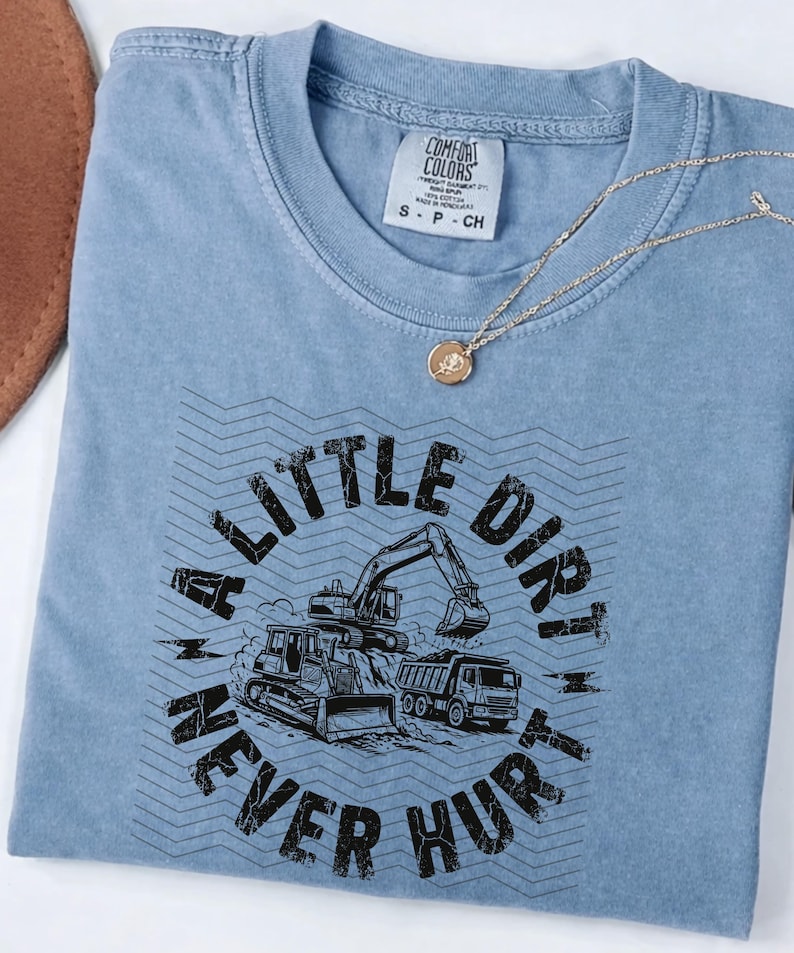 May include: A light blue t-shirt with the text "A Little Dirt Never Hurt" and illustrations of construction vehicles. A gold necklace with a circular pendant is on top of the shirt.