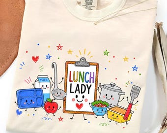 May include: A cream-colored t-shirt featuring a graphic print of anthropomorphic kitchen items including a clipboard labeled LUNCH LADY, a milk carton, a blue tray, an apple, a pot, a bowl of salad, and a spatula, surrounded by colorful stars.