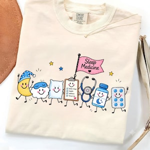 Peut inclure: Un t-shirt couleur crème avec un motif de personnages de dessins animés incluant un croissant de lune, un nuage, un oreiller, un presse-papiers, un stéthoscope, un flacon de médicament et une plaquette de pilules. Un drapeau rose indique Sleep Medicine.