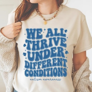 May include: Cream-colored t-shirt with the text "WE ALL THRIVE UNDER DIFFERENT CONDITIONS" in blue, with the words "autism awareness" below. The shirt has a crew neck and short sleeves.