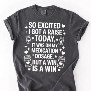 K&ouml;nnte beinhalten: Dunkelgraues T-Shirt mit wei&szlig;em Text: "SO EXCITED I GOT A RAISE TODAY, IT WAS ON MY MEDICATION DOSAGE, BUT A WIN IS A WIN." Das Design umfasst Pillenflaschen-Grafiken, Herzen und Sterne.