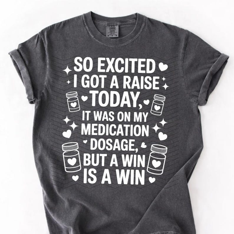 K&ouml;nnte beinhalten: Dunkelgraues T-Shirt mit wei&szlig;em Text: "SO EXCITED I GOT A RAISE TODAY, IT WAS ON MY MEDICATION DOSAGE, BUT A WIN IS A WIN." Das Design umfasst Pillenflaschen-Grafiken, Herzen und Sterne.