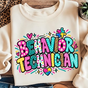 May include: A cream-colored crewneck sweatshirt featuring the text BEHAVIOR TECHNICIAN in large, multi-colored letters with various patterns including leopard print, daisies, and hearts, accented by colorful puzzle pieces and small hearts.