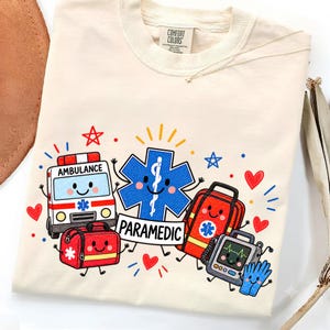 Könnte beinhalten: Ein cremefarbenes T-Shirt mit einem Grafikdruck von medizinischen Cartoon-Gegenständen, darunter ein Krankenwagen, ein blaues Stern-des-Lebens-Symbol mit dem Text PARAMEDIC, eine rote Arzttasche, ein Herzmonitor und Handschuhe, akzentuiert mit roten Herzen.