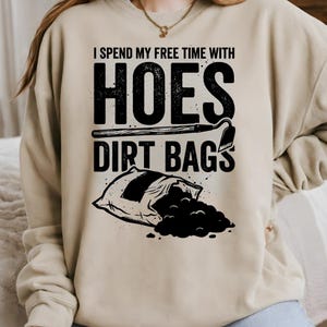 May include: Beige sweatshirt featuring the text "I spend my free time with hoes dirt bags" in black. The design includes a hoe and a bag of dirt. A gardening-themed garment.