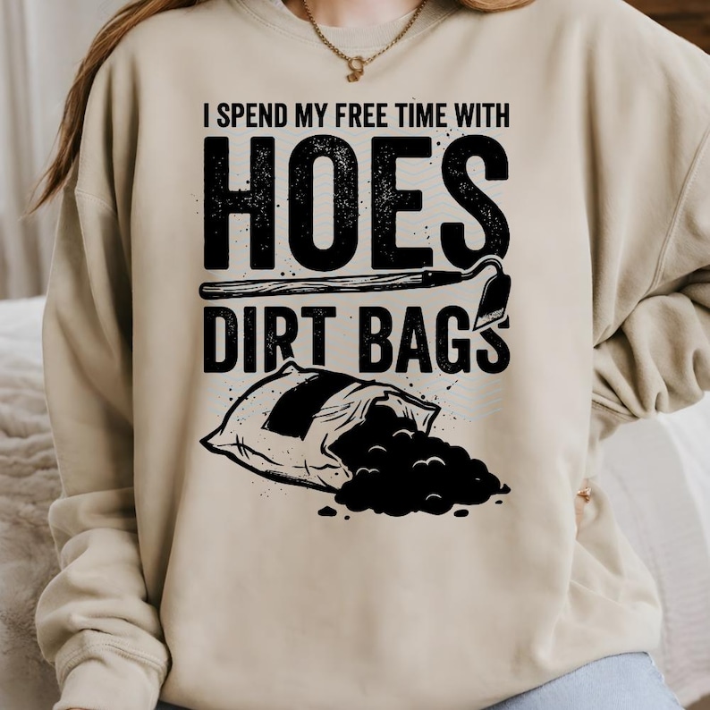 May include: Beige sweatshirt featuring the text "I spend my free time with hoes dirt bags" in black. The design includes a hoe and a bag of dirt. A gardening-themed garment.