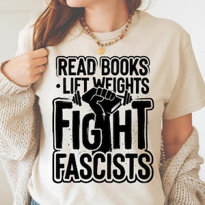 Puede incluir: Una camiseta color crema con el texto "READ BOOKS • LIFT WEIGHTS FIGHT FASCISTS" en negro. El diseño incluye un gráfico de una mano sosteniendo un libro con pesas. La camiseta es una prenda informal.