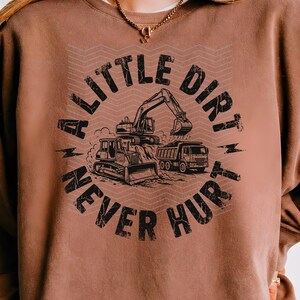 May include: Brown sweatshirt with a graphic design featuring construction vehicles: a bulldozer, excavator, and dump truck. The text "A LITTLE DIRT NEVER HURT" is printed in a distressed font, creating a vintage look.