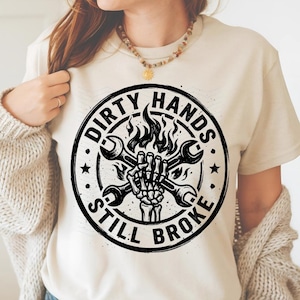 May include: Cream-colored t-shirt featuring a circular graphic with the text "DIRTY HANDS STILL BROKE." The design includes crossed wrenches, flames, and a skeleton hand. The graphic is in black and white.