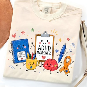 以下が含まれることがあります： クリーム色のTシャツで、文房具や脳のキャラクターが描かれたカラフルなグラフィックが特徴です。中央のクリップボードには「ADHD AWARENESS」の文字があり、青いノートには「FOCUS STRENGTHS POTENTIAL」と書かれています。星やハートの模様も含まれています。