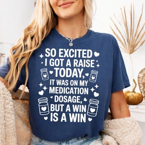 K&ouml;nnte beinhalten: Marineblaues T-Shirt mit wei&szlig;em Text: &bdquo;SO EXCITED I GOT A RAISE TODAY, IT WAS ON MY MEDICATION DOSAGE, BUT A WIN IS A WIN.&ldquo; Das Design umfasst Pillenflaschen-Grafiken und Sternakzente. Ein l&auml;ssiges, humorvolles T-Shirt.
