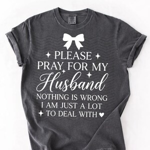 May include: Dark grey t-shirt with white text: "PLEASE PRAY FOR MY Husband NOTHING IS WRONG I AM JUST A LOT TO DEAL WITH." Features a white bow graphic at the top.