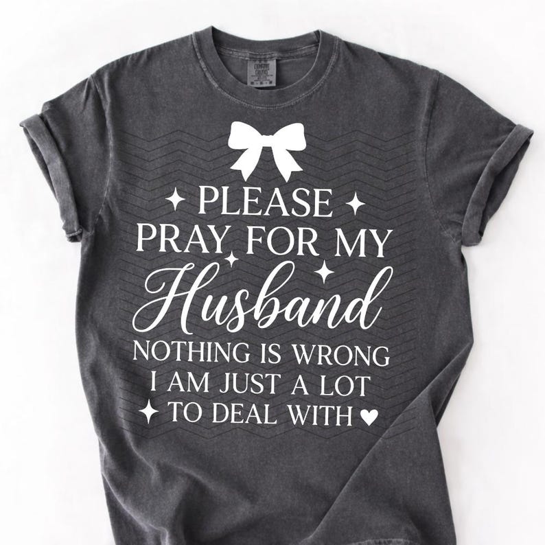 May include: Dark grey t-shirt with white text: "PLEASE PRAY FOR MY Husband NOTHING IS WRONG I AM JUST A LOT TO DEAL WITH." Features a white bow graphic at the top.