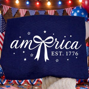 May include: A dark blue t-shirt featuring white text that reads am-rica with a white bow replacing the letter e. Below the text, it says EST. 1776. The design includes several small white stars scattered around the text and a subtle chevron pattern.